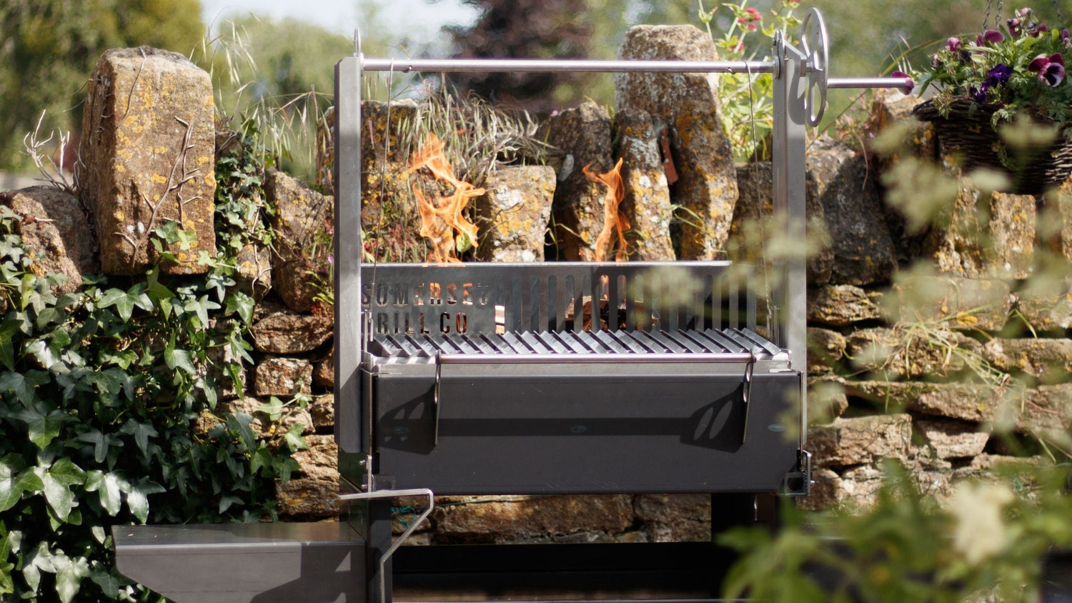 Stainless steel wood-fired grill with flames, set outdoors against a rustic stone wall