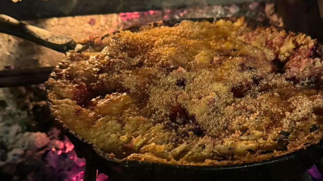 Golden brown apple and blackberry crumbly pudding cooking over glowing embers in a cast iron skillet