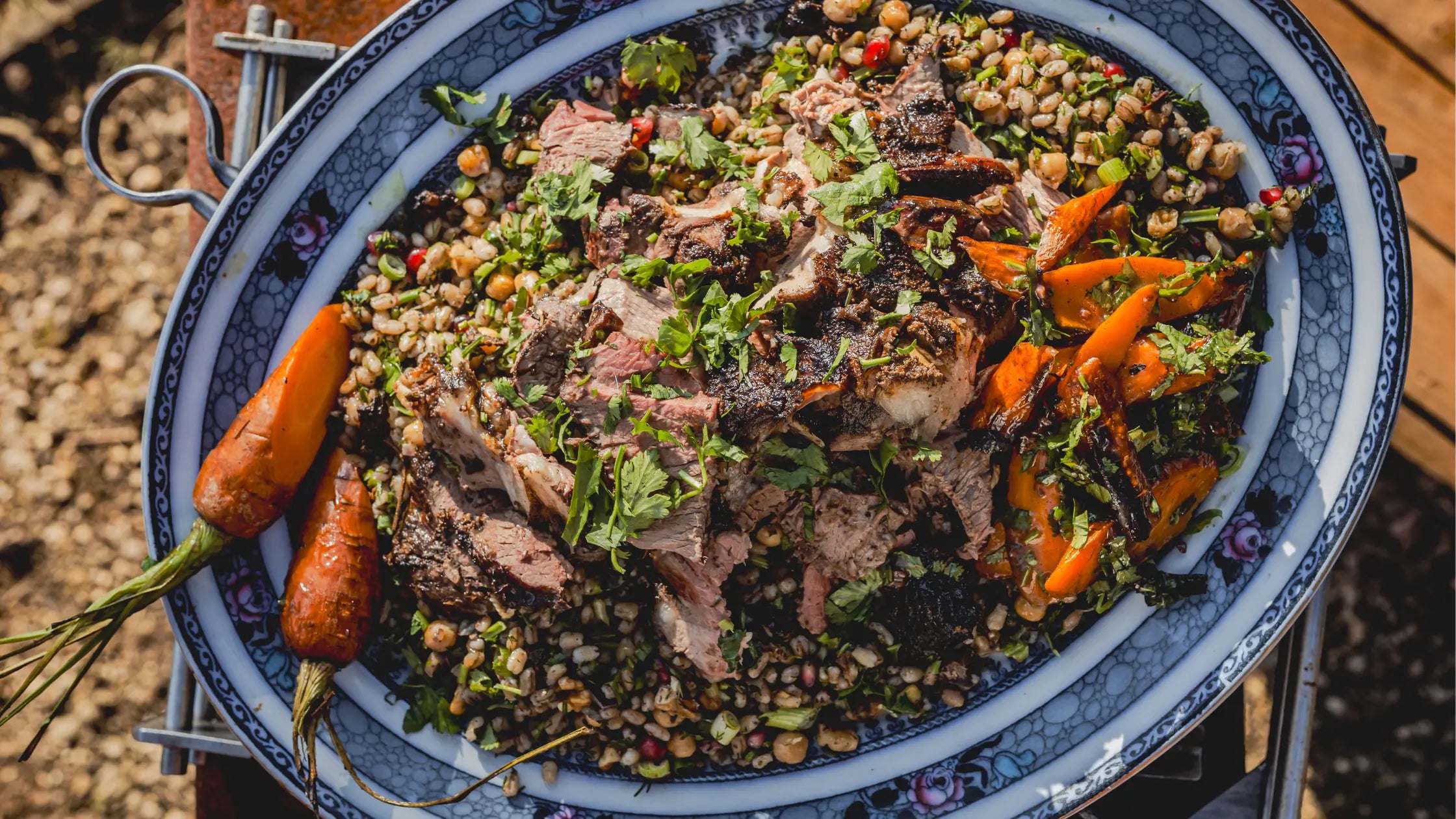 Harissa-spiced rotisserie lamb served on a blue patterned platter with grilled carrots and mixed grain salad garnished with fresh herbs