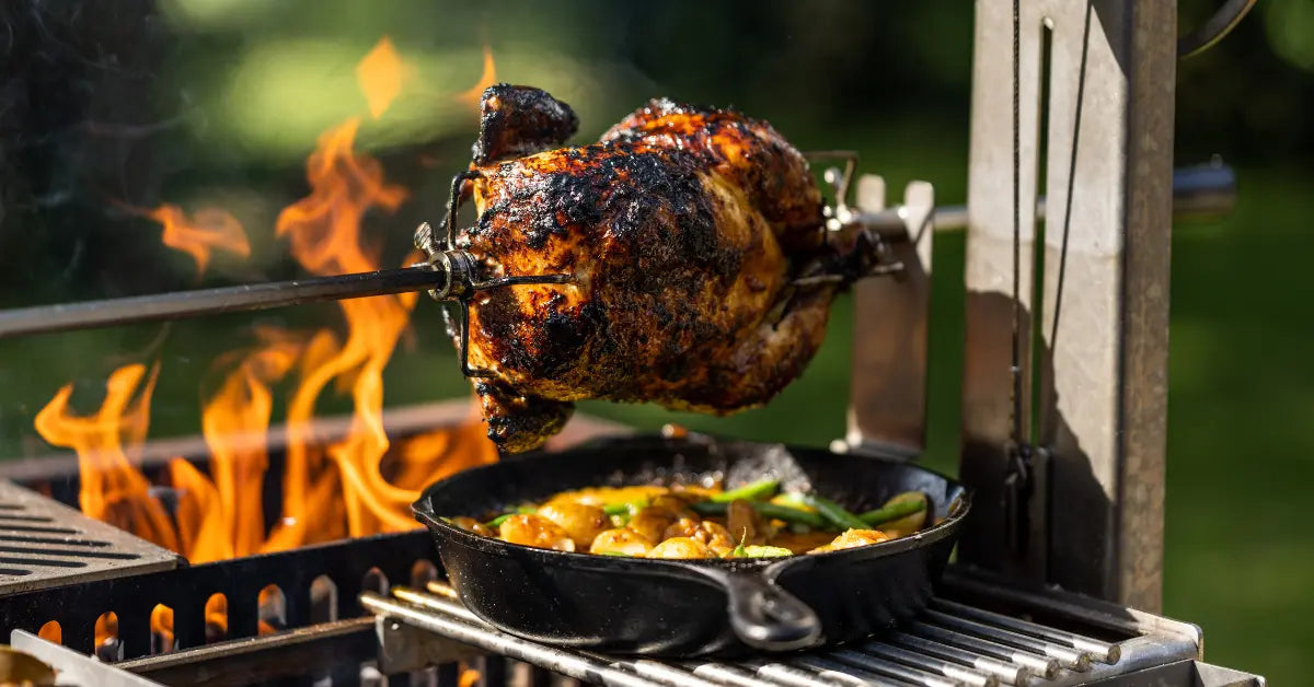 Whole chicken roasting on a spit over open flames with vegetables cooking in a cast iron pan below