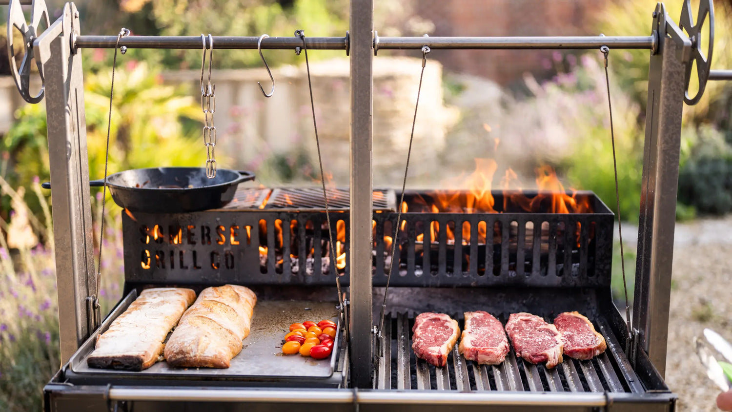 Traditional Argentinian grill with flames, four steaks on grill, bread and cherry tomatoes on flat surface
