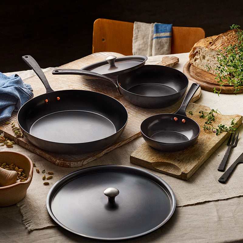 Set of black cast iron frying pans and lids on wooden boards with bread and herbs nearby