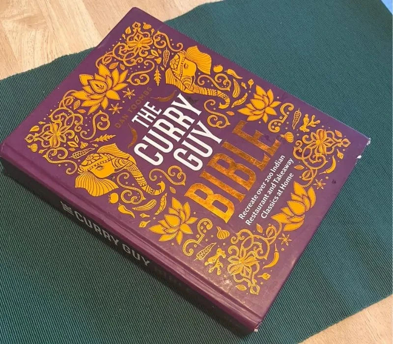 Purple hardcover cookbook titled The Curry Guy Bible with intricate orange floral and elephant designs