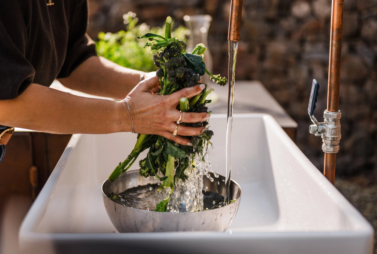 A person washing a bunch of green broccoli in a metal bowl set in a white sink, with water running from copper pipes in a rustic kitchen setting.