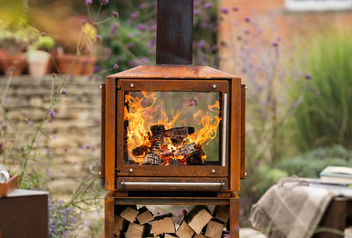 Close-up of the fire burning behind the Cor-Ten Outdoor Woodburner's glass, with logs stacked in the log store below
