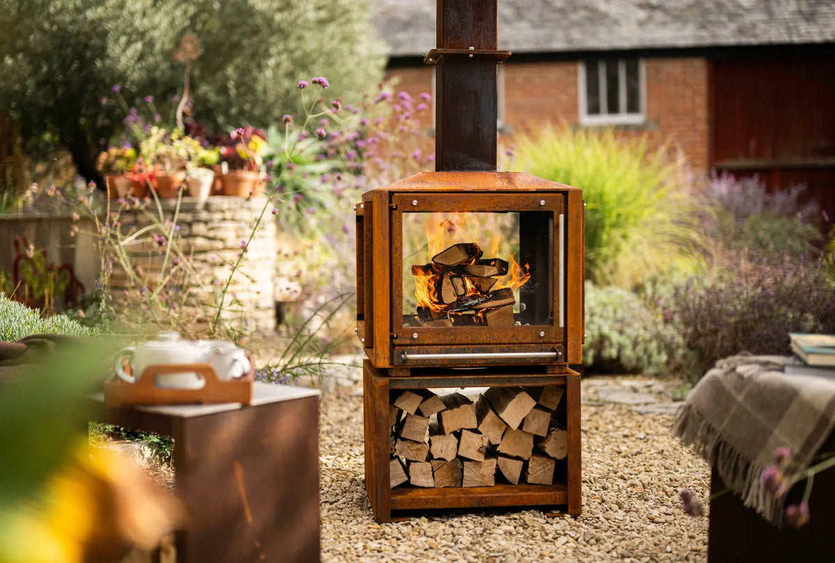 Cor-Ten Outdoor Woodburner alight in a garden, with its log store full and a Cor-Ten bench alongside