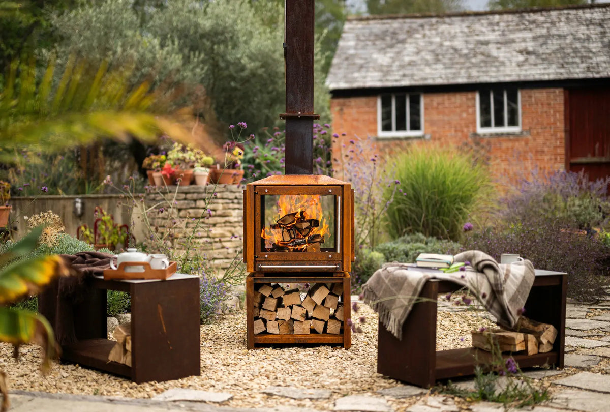 Cor-Ten Outdoor Woodburner alight between two Cor-Ten benches, one with a tea tray and one with a blanket