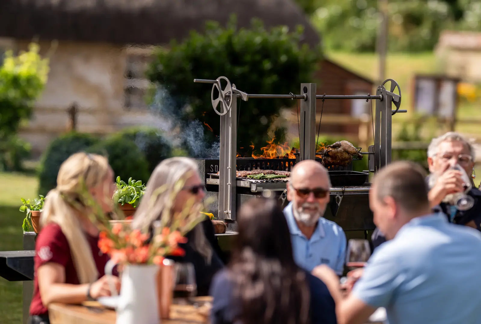 Gunmetal grey outdoor kitchen grill with rotisserie cooking chicken and vegetables, people dining outdoors