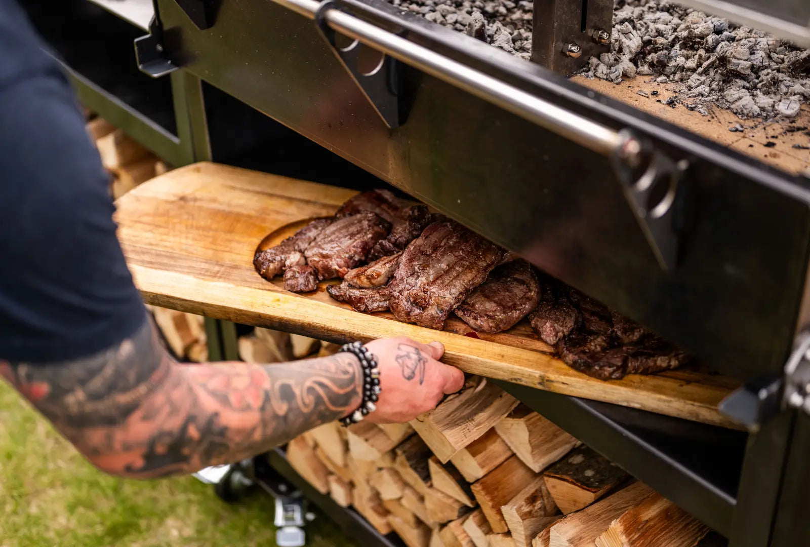 Tattooed arm placing grilled steaks on wooden board inside gunmetal outdoor kitchen with stacked firewood