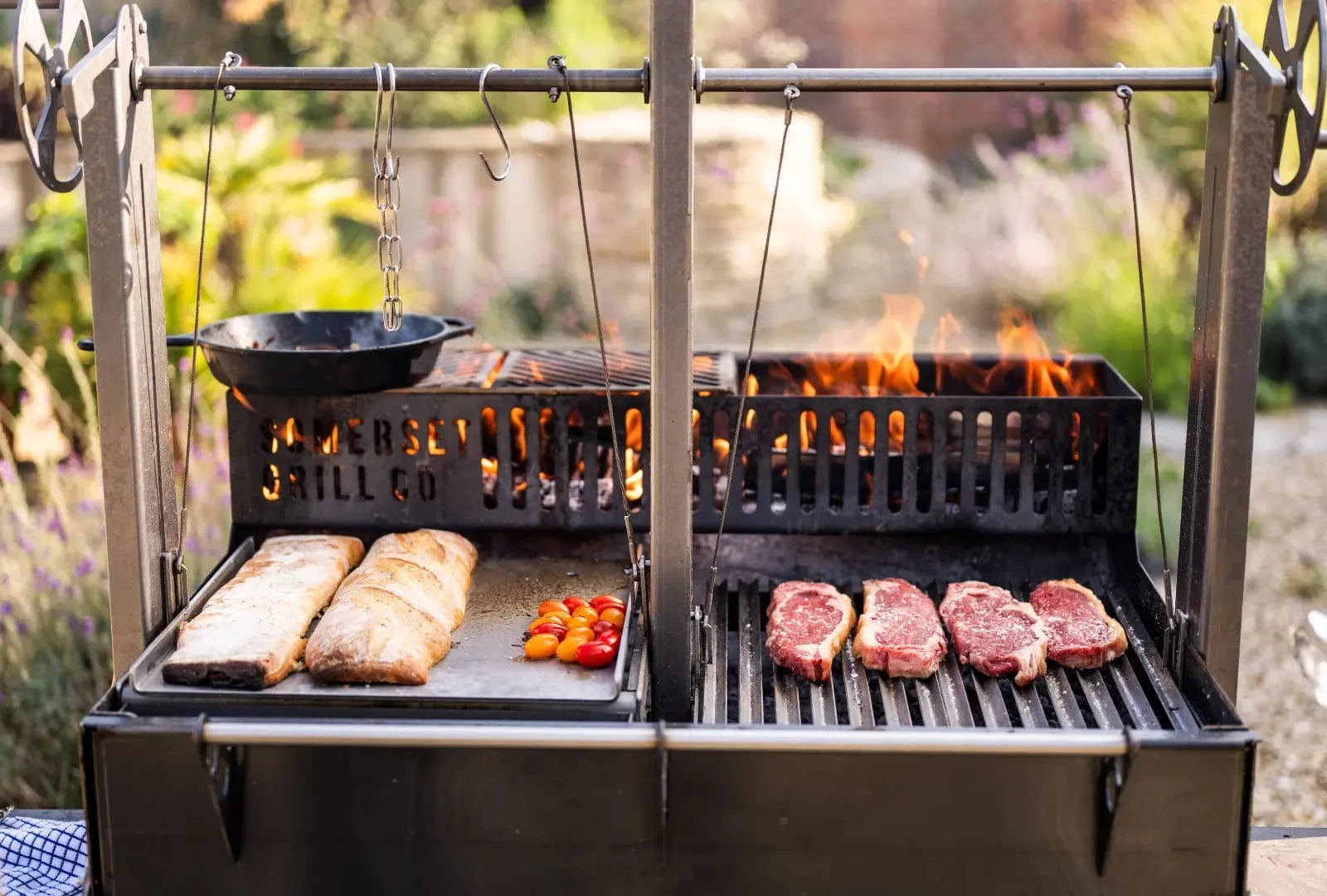 Gunmetal outdoor kitchen bundle with grill, griddle, hanging cast iron pan, bread, tomatoes, and steaks cooking
