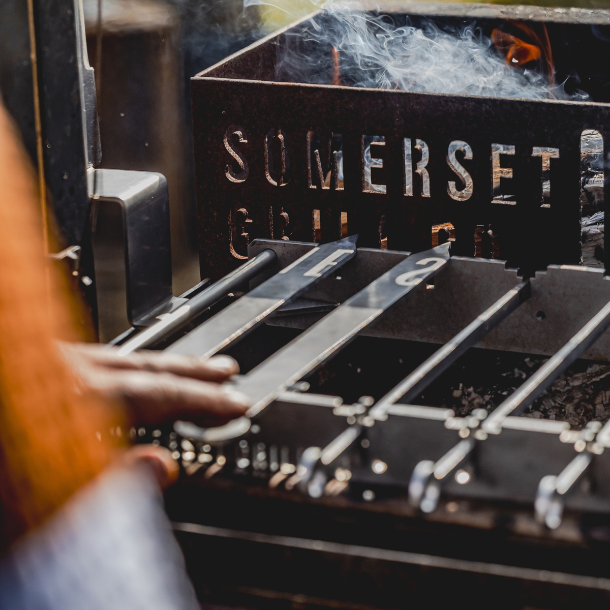 Close-up of a hand adjusting metal grill grates in front of a black metal smoker box with the brand name SOMERSET GRILL visible, with smoke rising and a blurred outdoor background.