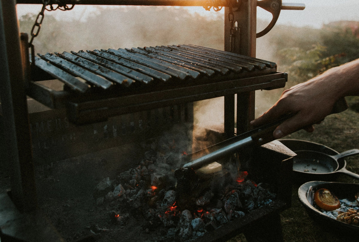 A close-up outdoor shot of a hand using tongs to adjust coals under a smoking grill . Warm sunlight and smoke fill the background, with cooking pans visible on the side.