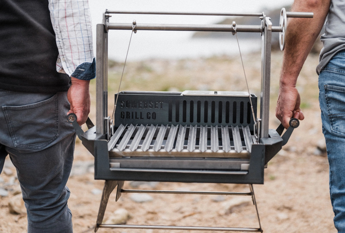 A person holds an Asado Mini grill outdoors on a sandy ground, with another person partially visible on the right side.