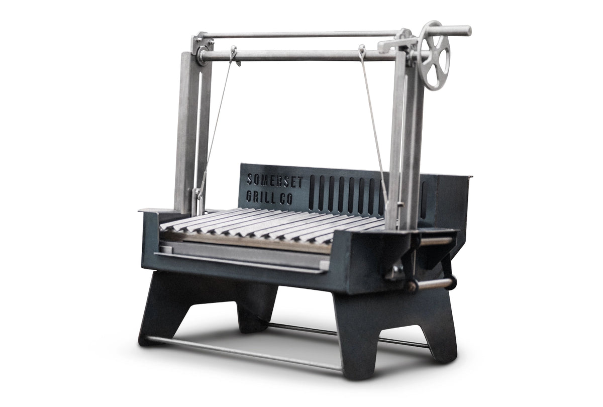 An Asado Mini grill with a hand-crank wheel on top, featuring a dark gray cast iron body and silver metal grate, shown on a white background.