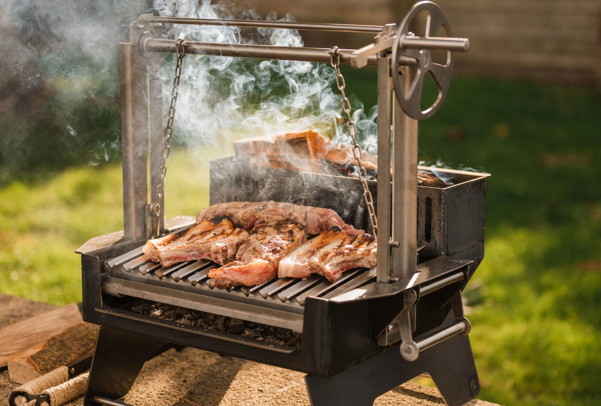 An Asado Mini grill with a wheel handle, shown outdoors with meat cooking on the grate and smoke rising against a blurred green lawn background.