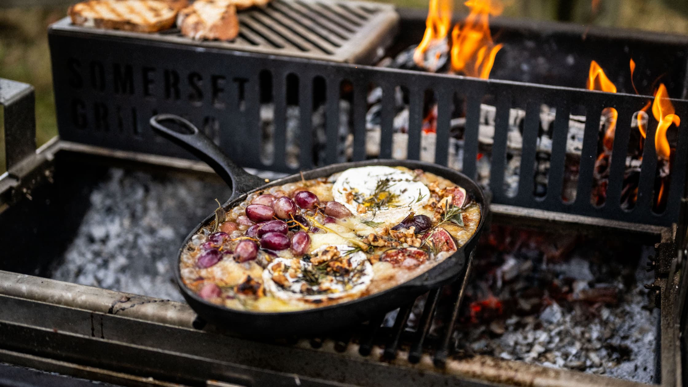 Cast iron skillet with melted cheese, grapes, walnuts, and herbs cooking on outdoor grill
