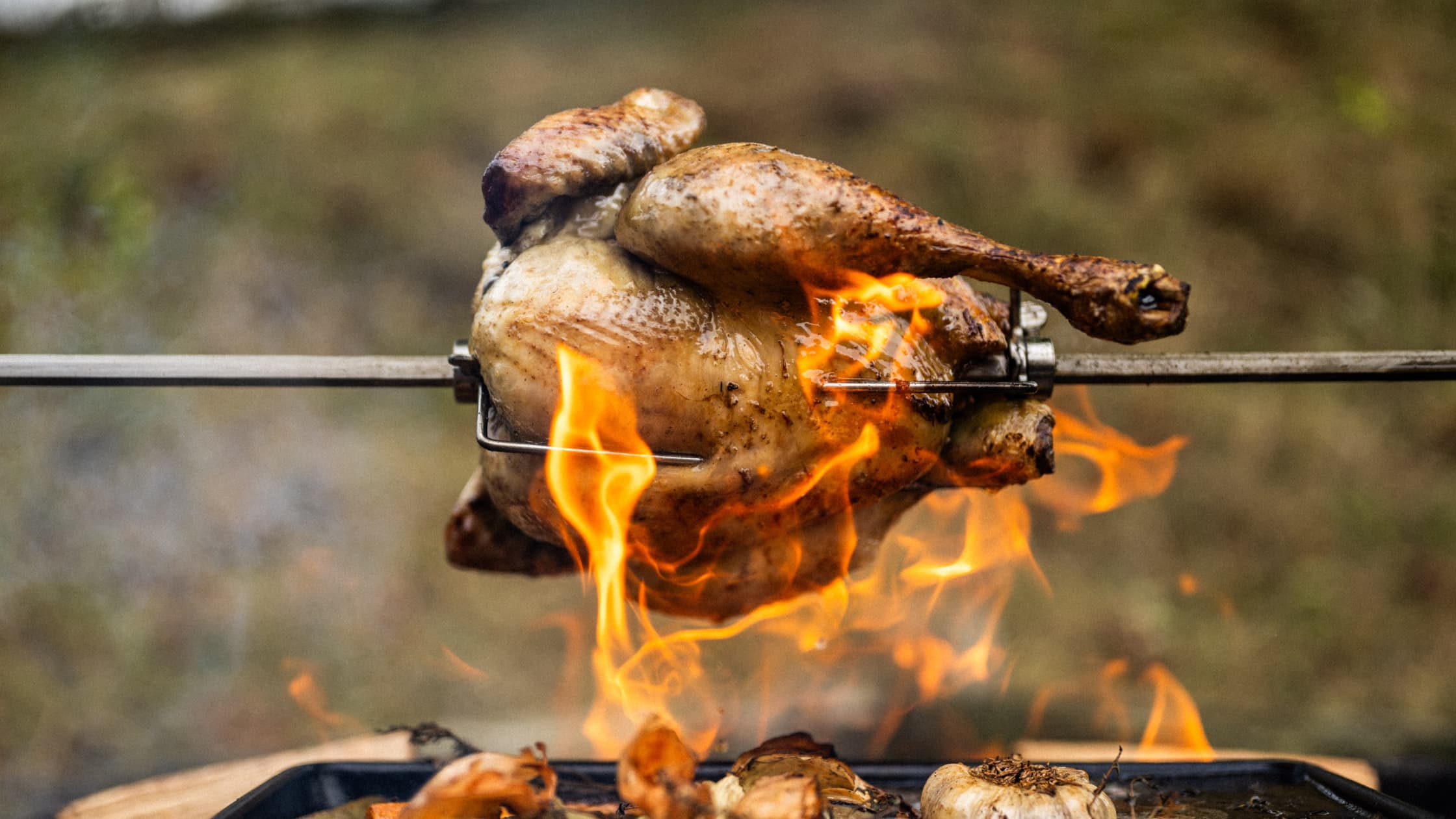 Whole rotisserie chicken roasting over open flames on a metal spit outdoors