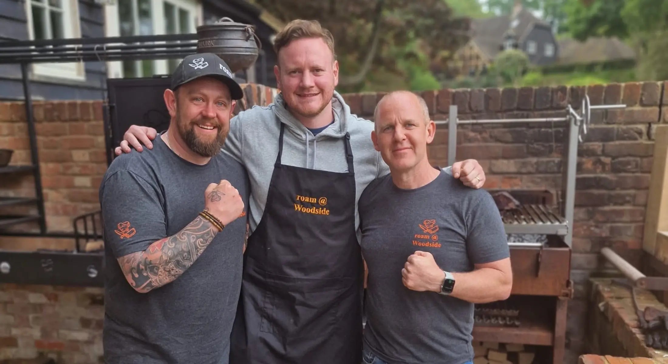 Three men smiling outdoors by brick wall, two wearing grey t-shirts and one black apron with cooking school logo