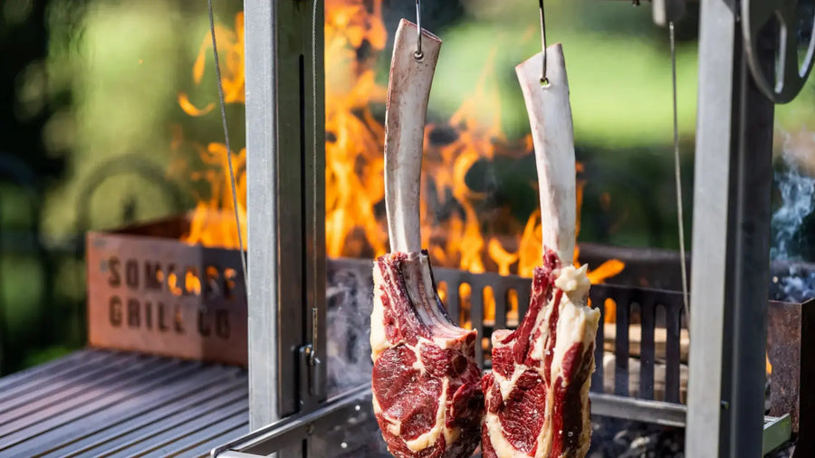 Two large raw tomahawk steaks hanging on hooks over an open flame grill outdoors
