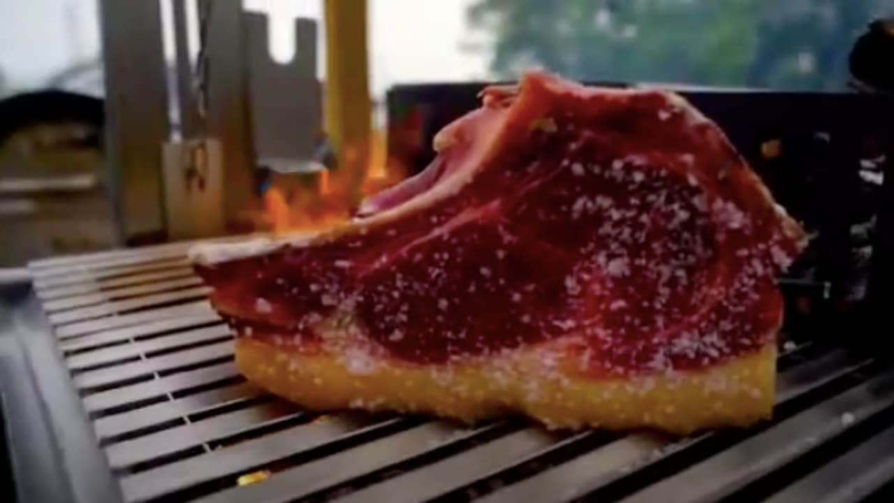 Bone-in beef sirloin steak seasoned with salt cooking on a metal grill over open flame