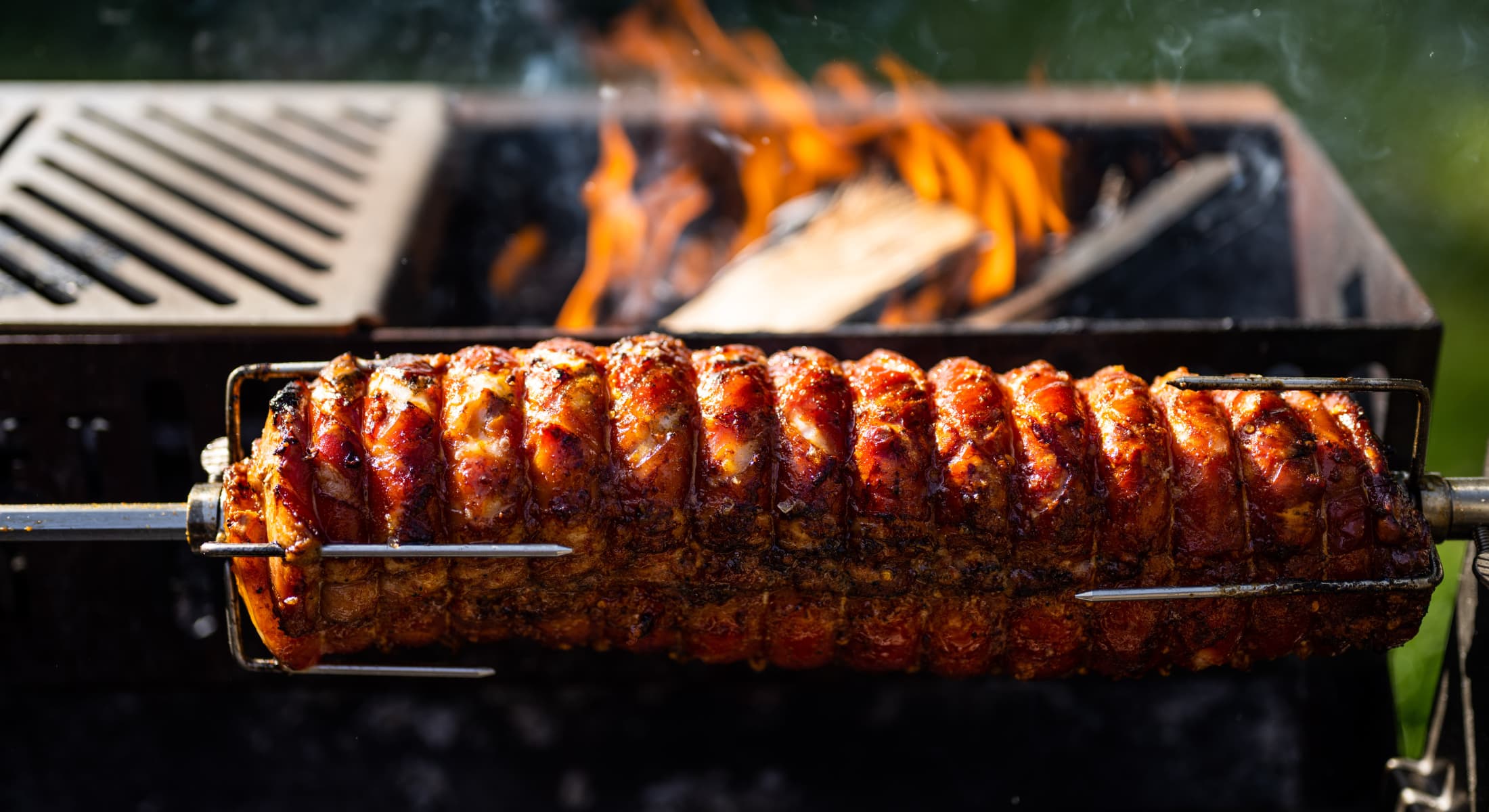Golden-brown nduja stuffed porchetta roasting on a spit over an open flame grill