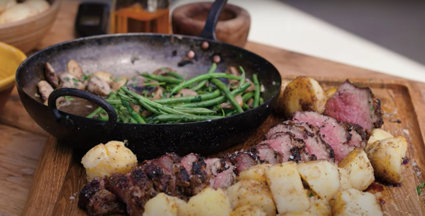 Sliced Santa Maria style tri-tip steak with roasted potatoes and green beans in a black skillet on wooden board
