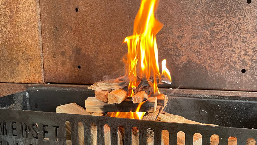 Bright orange flames burning small wooden logs in a black metal fire grate with rusty background