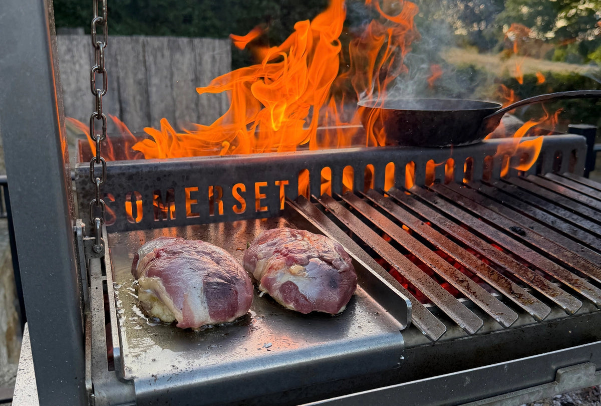 Duck cooking on a Somerset outdoor grill with bright orange flames and smoke, set against a blurred backyard background.