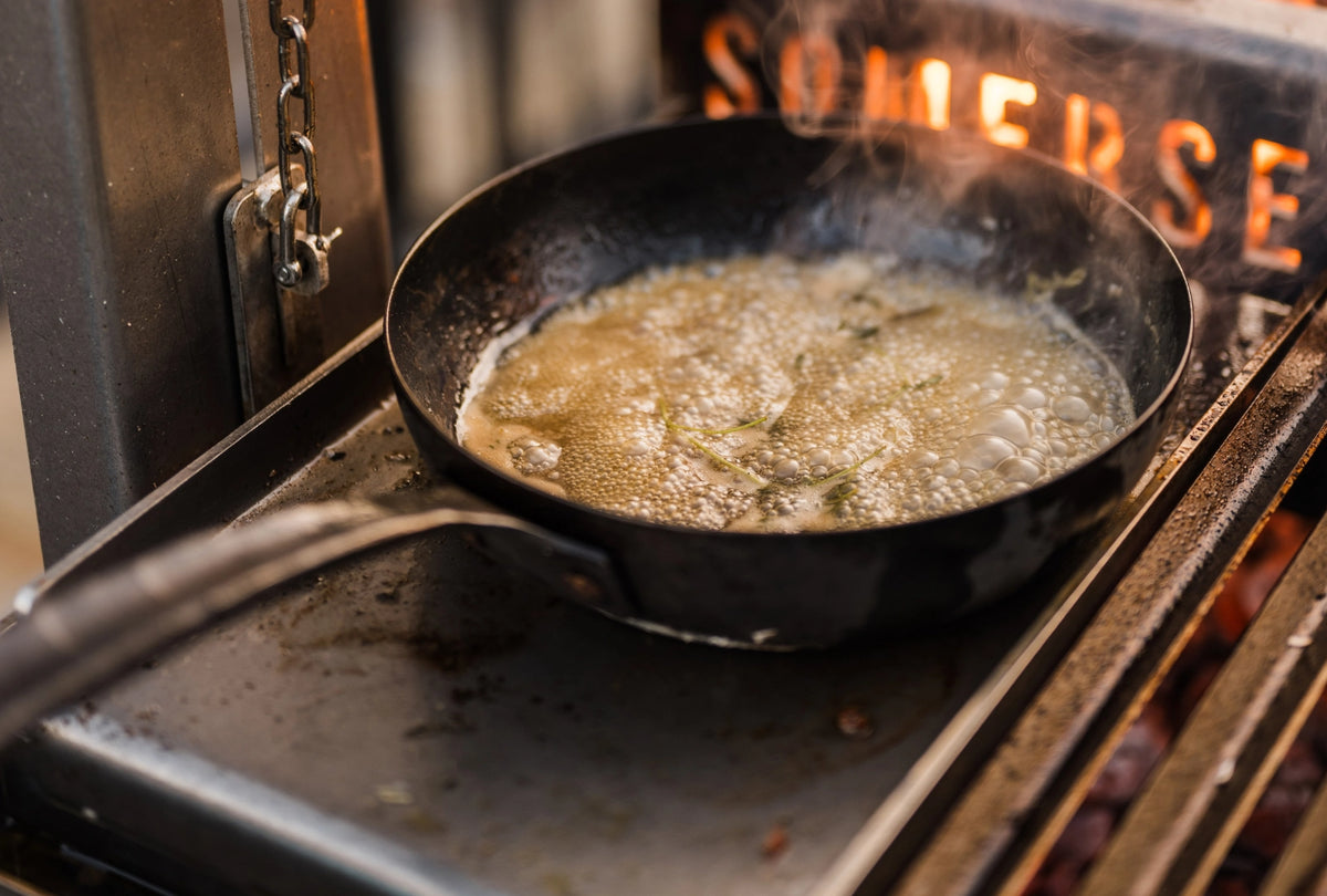 A black cast iron skillet with a long handle sits on a chapa, cooking a bubbling light brown sauce or butter with herbs. 