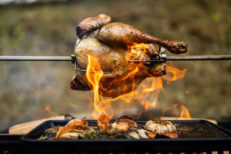 Whole chicken roasting on a rotisserie spit over open flames with vegetables below