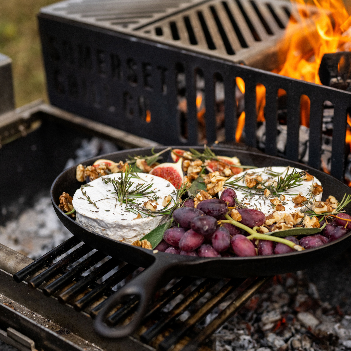 A cast iron skillet sits on a lit grill over coals, cooking a cheese and fig spread with nuts, grapes, and rosemary. A black cast iron grill grate with the words 'SOMERSET GRILLING' is visible in the background above an open flame.