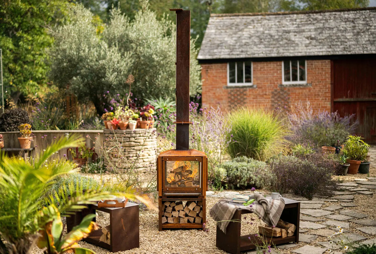 Cor-Ten Outdoor Woodburner lit on a gravel garden terrace, logs stacked in the log store beneath, between two Cor-Ten benches