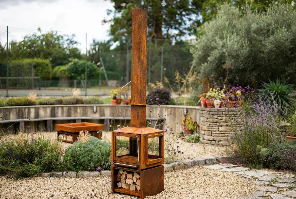 Cor-Ten Outdoor Woodburner with its tall chimney and a full log store, in a gravel garden