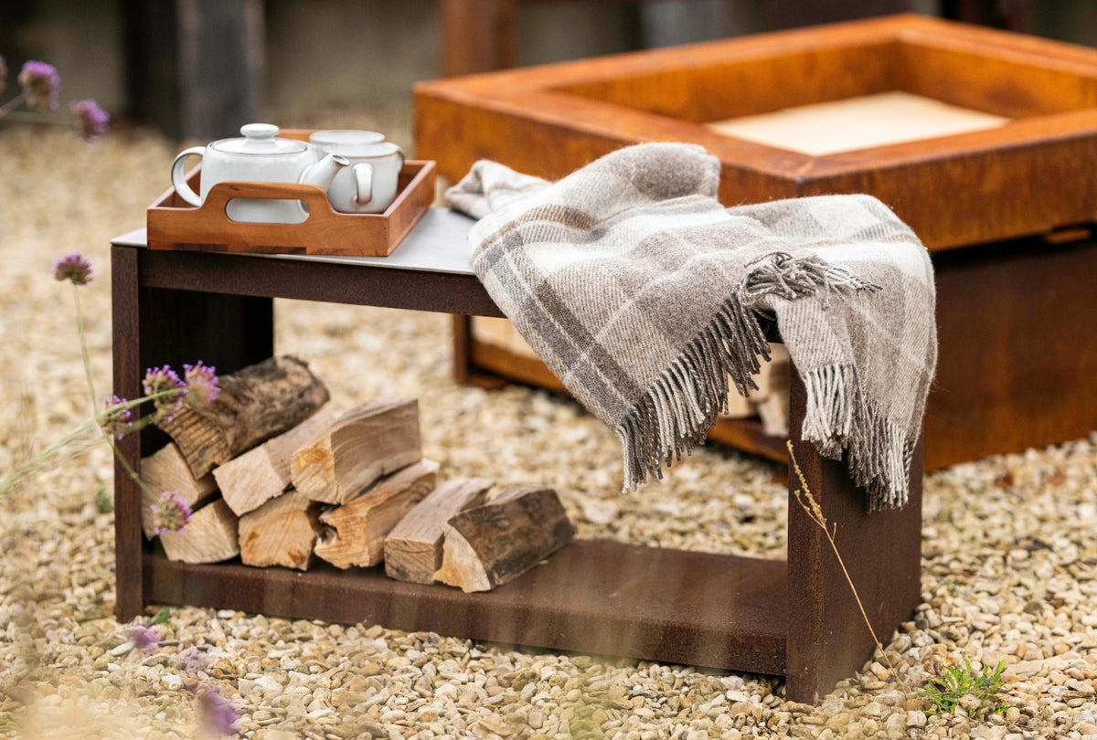 A rustic Cor-Ten wood storage bench or table styled outdoors on a gravel surface, holding a white teapot in a wooden tray, a beige plaid throw blanket, and stacked firewood in the lower shelf.