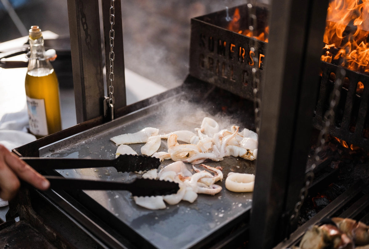 Squid and calamari cooking on a chapa, with flames visible in the background and a bottle of oil on the side.