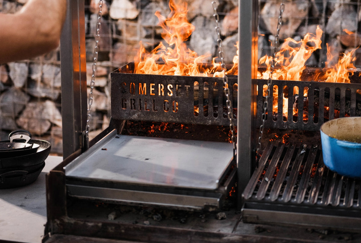 A close-up of a Grande grill with visible flames, a Chapa, and a blue pot on the right side. A person's arm is visible on the left, and a stone wall is in the background.