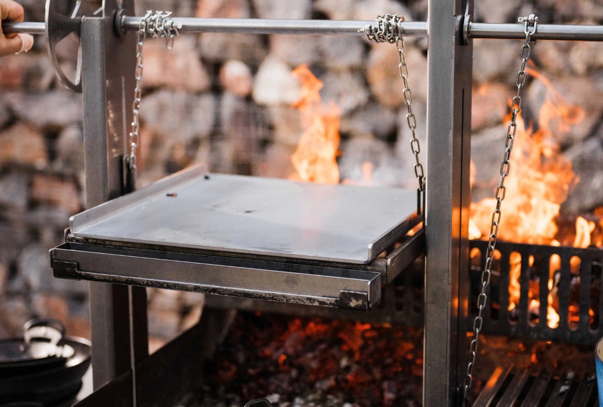 A chapa suspended by chains from a horizontal bar, positioned over an open fire with visible flames and embers in the background.