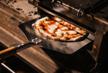 A freshly baked pizza with melted cheese and red sauce sits on a metal peel inside a rustic wood-fired oven.