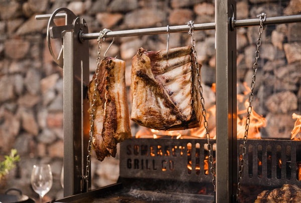 Two large racks of ribs hanging from metal hooks on a vertical grill frame, cooking over visible flames with a blurred stone wall background.