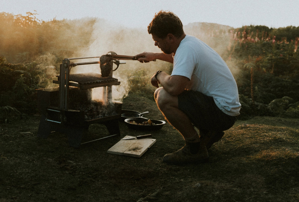 A man crouches outdoors at sunset next to a smoking portable grill, holding a pan over the fire. He wears a white t-shirt and dark shorts in a grassy field with trees in the background.