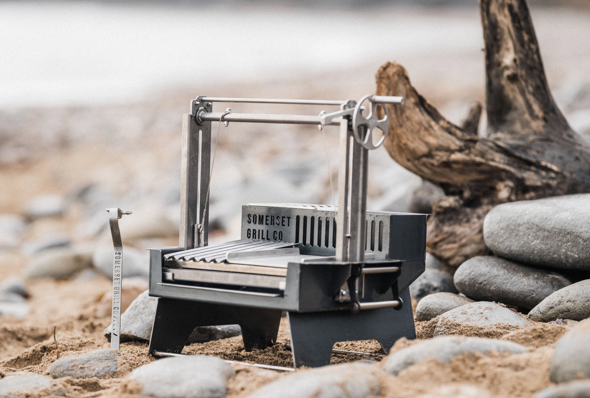 An Adsado Mini Grill Someset Grill Co. tabletop fire pit and grill sits on a sandy, rocky beach next to driftwood.y 