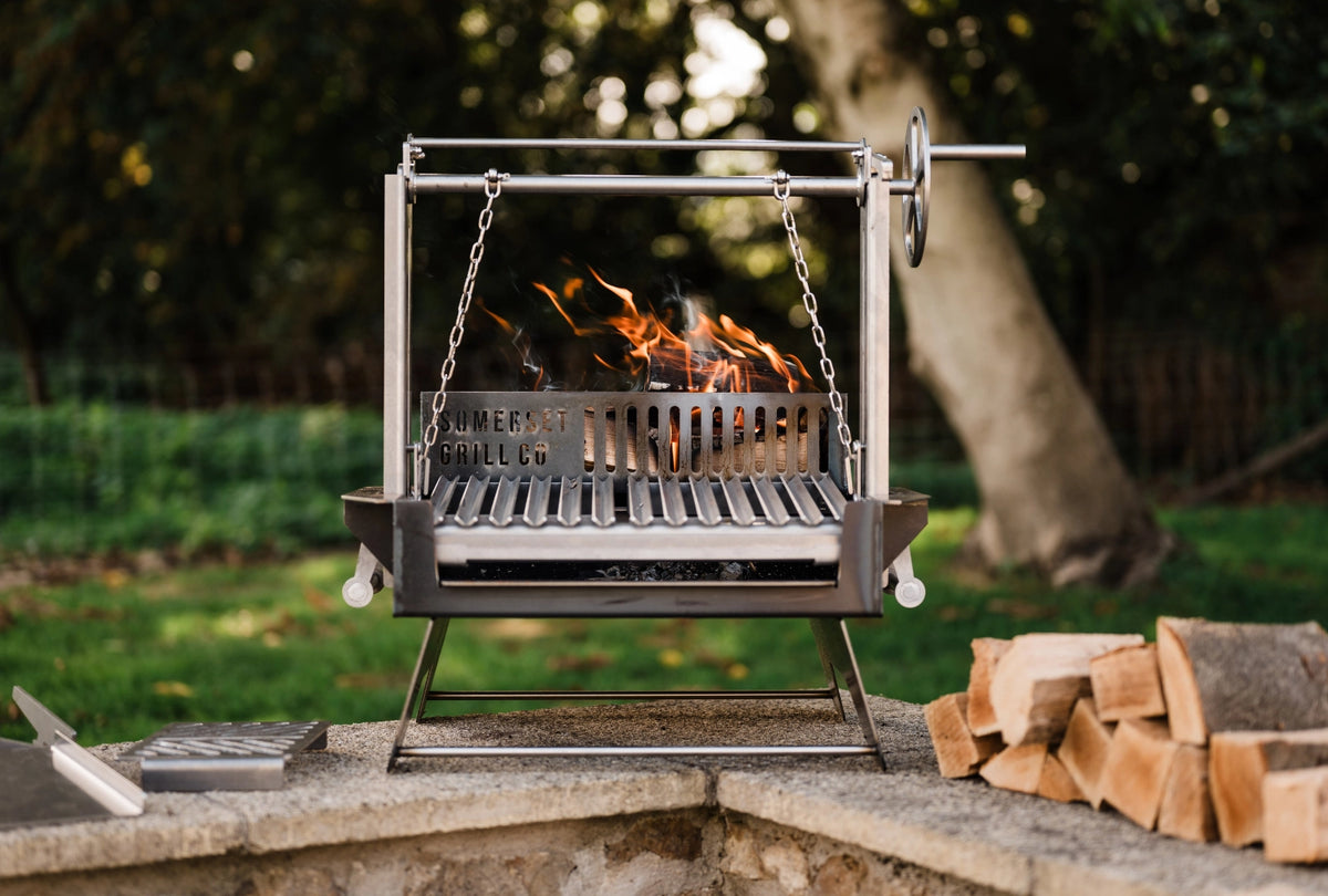 An Asado Mini steel outdoor grill with a visible fire, set on a stone patio with stacked firewood and a blurred garden background.