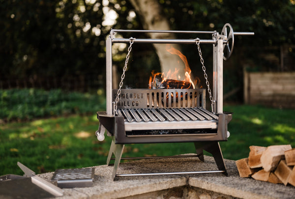 A Asado Mini grill with a hanging grate and visible flames, set outdoors on a stone surface with a blurred garden background and stacked firewood nearby.