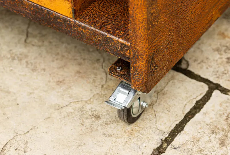 Close-up of Cor-Ten steel Classic Kamado Joe unit corner with metal caster wheel on stone floor
